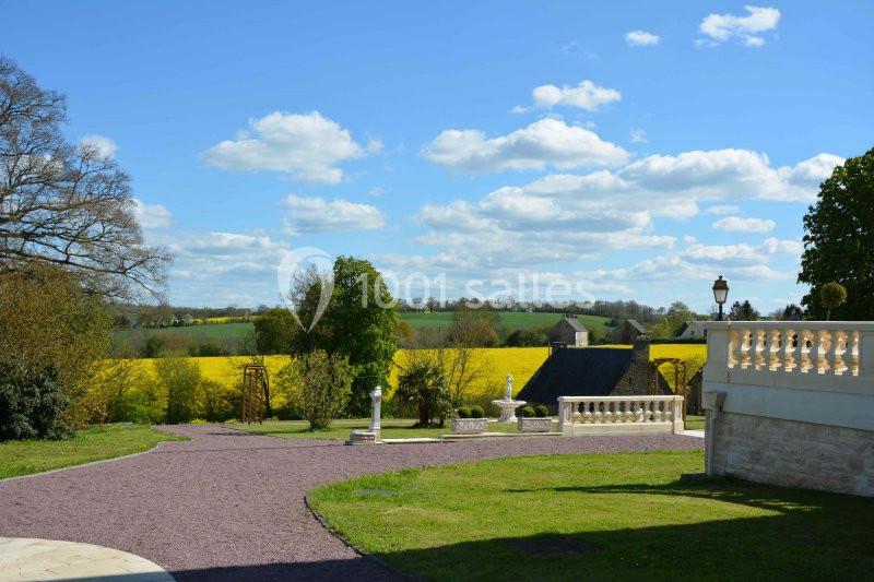Paysage rural avec un chemin en gravier, des champs de colza jaunes, des arbres et une terrasse en pierre au premier plan.