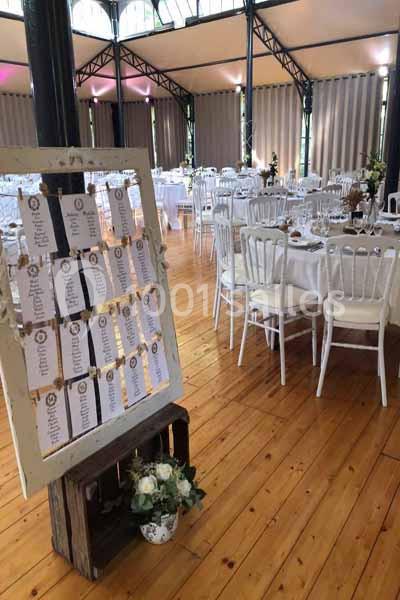 Plan de table sur chevalet dans une salle de réception lumineuse avec tables rondes dressées et chaises blanches.