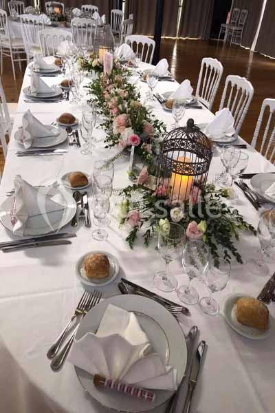 Table de réception élégamment dressée avec vaisselle blanche, décor floral et lanternes au centre.