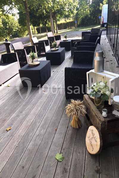 Terrasse en bois avec mobilier en rotin noir, décorée de plantes, épis de blé et lanternes, entourée de verdure.