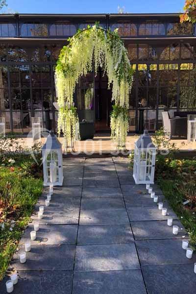 Allée en pierre bordée de bougies menant à une arche fleurie devant une verrière.