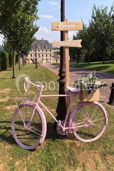 Vélo rose avec panier fleuri posé près d'un panneau en bois indiquant des directions, devant un chemin et un bâtiment.