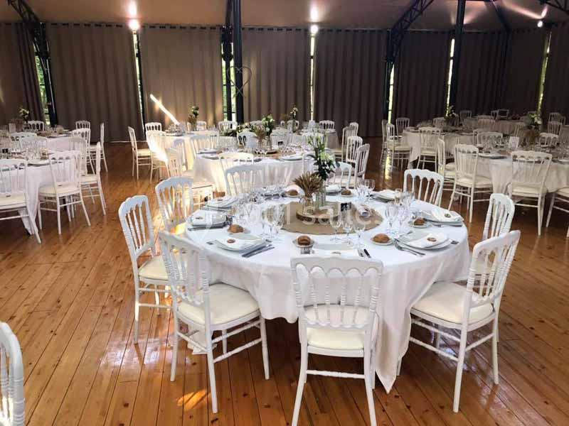 Salle de réception avec tables rondes dressées, nappes blanches, chaises en bois blanc et lumière naturelle.