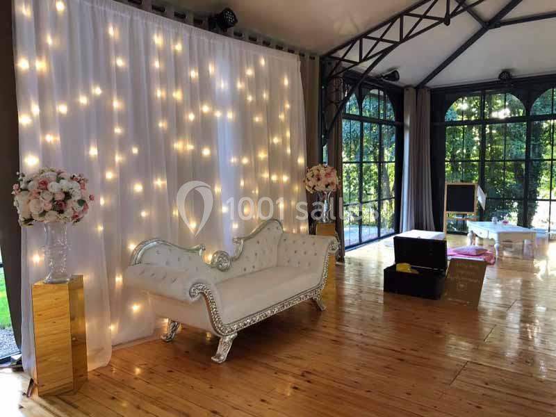 Canapé blanc capitonné devant un rideau lumineux, décoré de fleurs, dans une salle avec parquet et grandes baies vitrées.