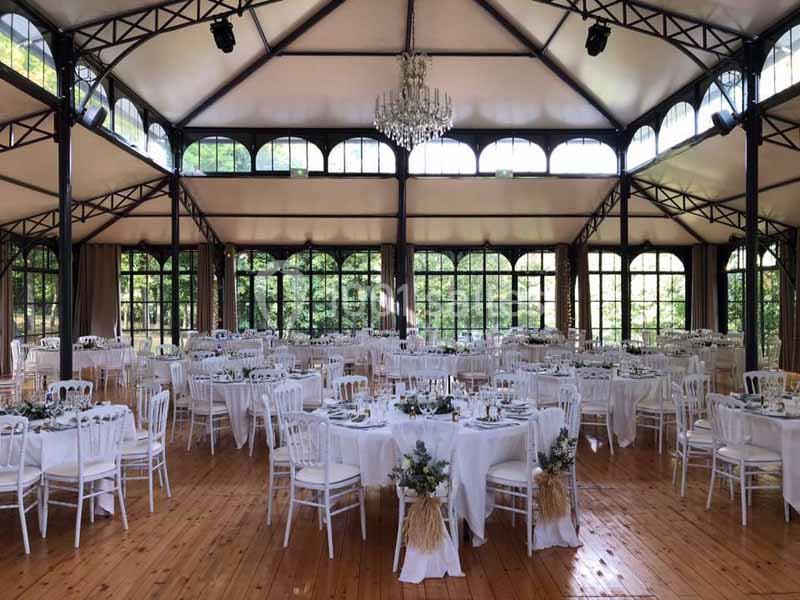 Salle de réception lumineuse avec tables rondes dressées, chaises blanches et un lustre central sous une verrière.