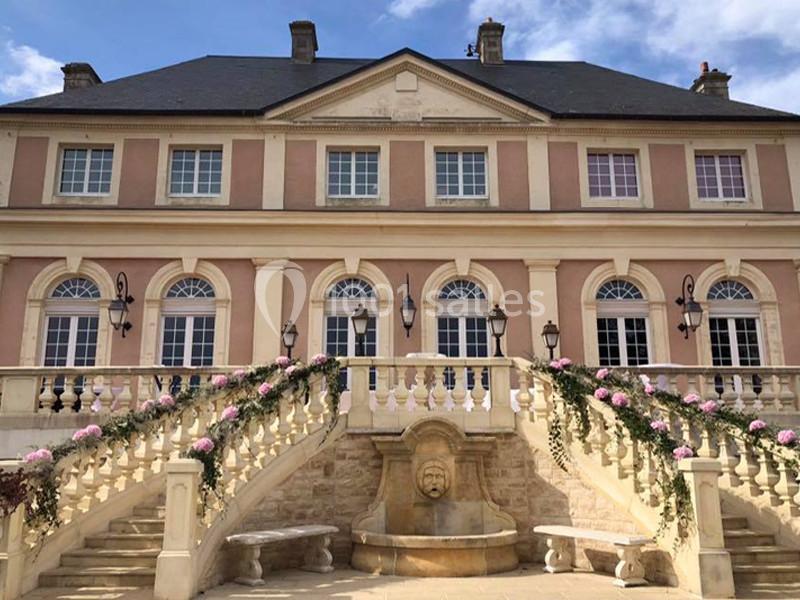 Façade d'un bâtiment élégant avec escalier symétrique, balustrades fleuries et fontaine ornée d'un visage sculpté.