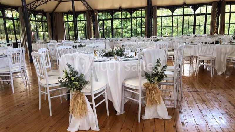 Salle de réception décorée avec des tables rondes, nappes blanches et chaises ornées de bouquets végétaux.