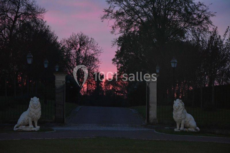 Deux statues de lions encadrent une allée bordée d'arbres, avec un ciel rose et violet au coucher du soleil.