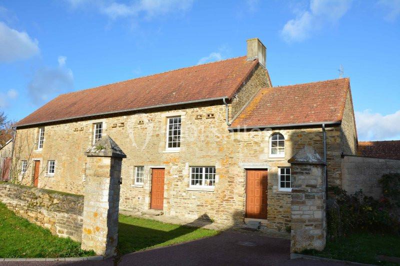 Maison en pierre avec toit en tuiles rouges, trois portes en bois et un jardin devant, sous un ciel bleu.