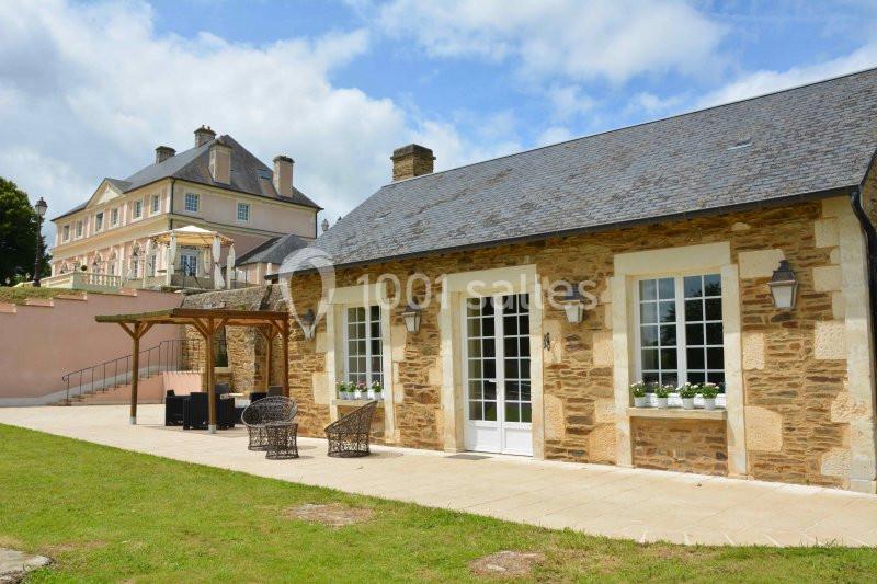 Bâtiment en pierre avec terrasse aménagée, situé devant une grande maison de style classique sous un ciel partiellement…