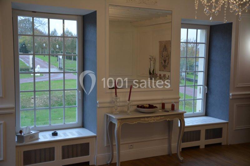Intérieur lumineux avec deux grandes fenêtres donnant sur un jardin, table décorée de bougies et miroir central.