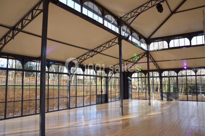 Salle lumineuse avec structure métallique, grandes baies vitrées et parquet en bois, donnant sur un extérieur arboré.