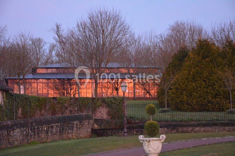 Bâtiment vitré illuminé au crépuscule, entouré d'arbres dénudés et d'un jardin avec une allée et une vasque décorative.