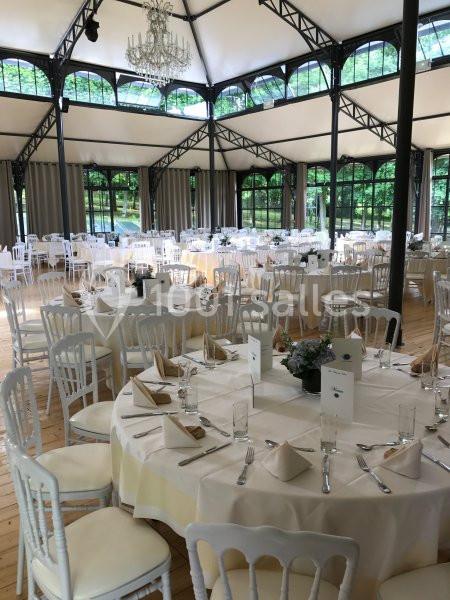Salle de réception lumineuse avec tables rondes dressées, nappes blanches et chaises en bois clair.