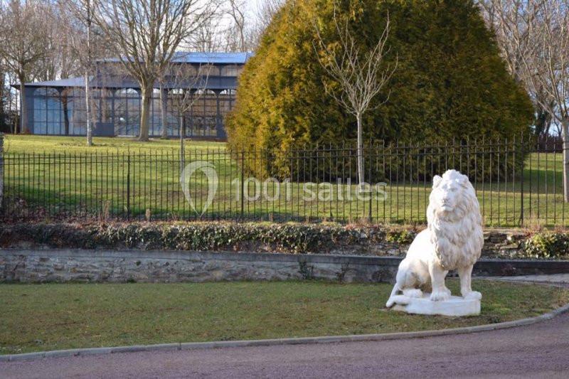 Statue de lion blanc posée sur une pelouse devant une clôture, avec des arbres et un bâtiment vitré en arrière-plan.