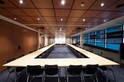 Salle de réunion avec une grande table en bois, chaises noires, parois vitrées et vue sur des immeubles.