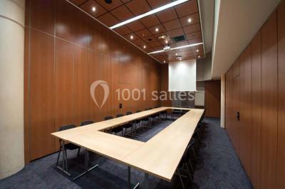 Salle de réunion avec une grande table en bois, chaises noires, parois vitrées et vue sur des immeubles.