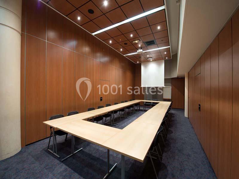 Salle de réunion vide avec tables disposées en U, chaises noires, murs en bois et éclairage au plafond.