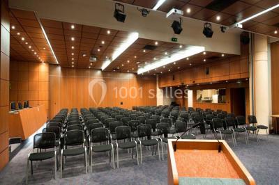 Salle de réunion avec une grande table en bois, chaises noires, parois vitrées et vue sur des immeubles.