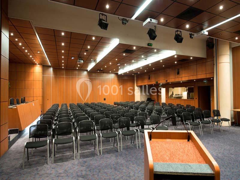 Salle de conférence vide avec rangées de chaises noires, pupitre au premier plan et éclairage au plafond.