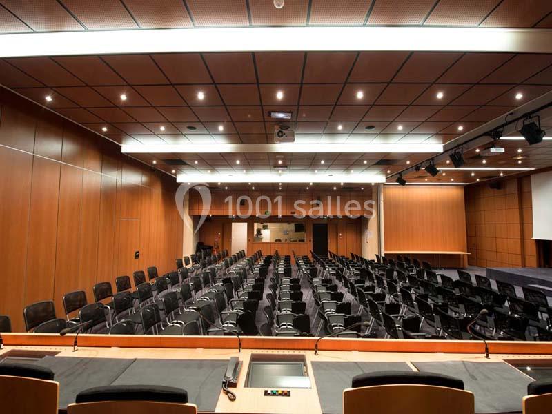 Salle de conférence vide avec rangées de sièges noirs, pupitres en bois et éclairage au plafond.