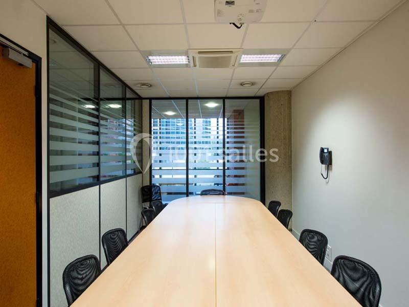 Salle de réunion avec une grande table en bois, chaises noires, parois vitrées et vue sur des immeubles.