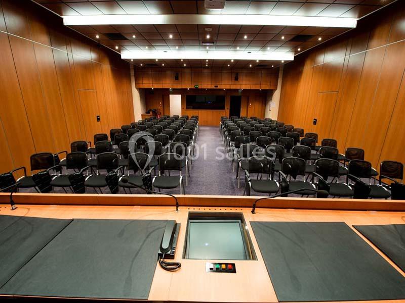 Salle de conférence vide avec rangées de chaises noires, vue depuis un pupitre équipé de commandes.