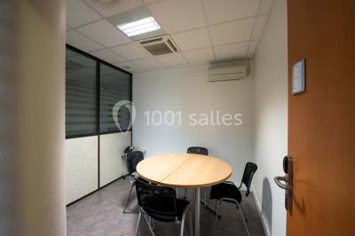 Salle de réunion avec une grande table en bois, chaises noires, parois vitrées et vue sur des immeubles.