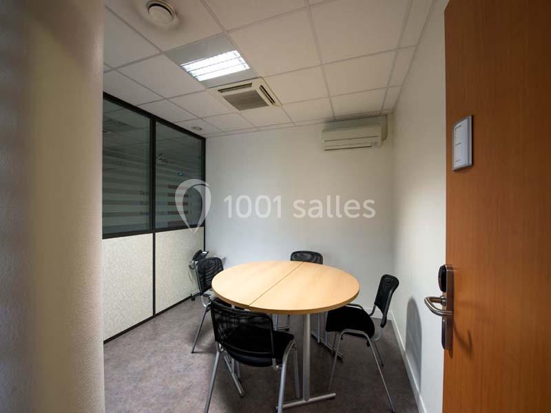 Salle de réunion compacte avec une table ronde, quatre chaises noires, murs blancs et éclairage au plafond.