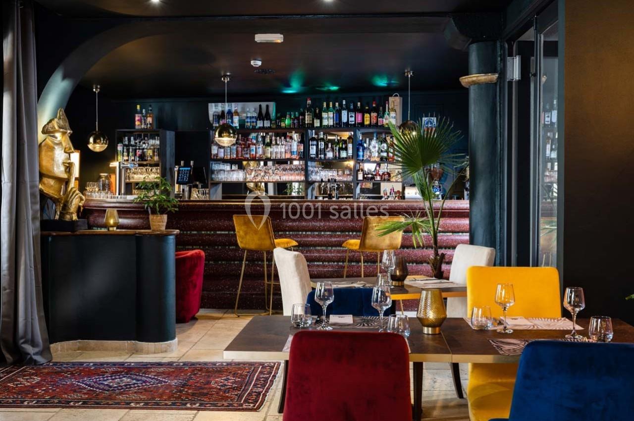 Intérieur d'un bar-restaurant élégant avec un comptoir en bois, des tables dressées et des fauteuils colorés.