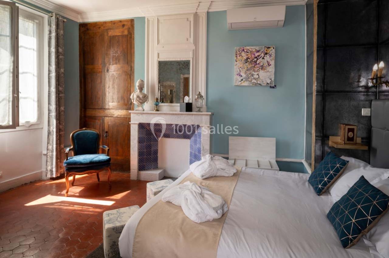 Chambre lumineuse avec lit double, fauteuil bleu, cheminée décorative et murs bleu clair.