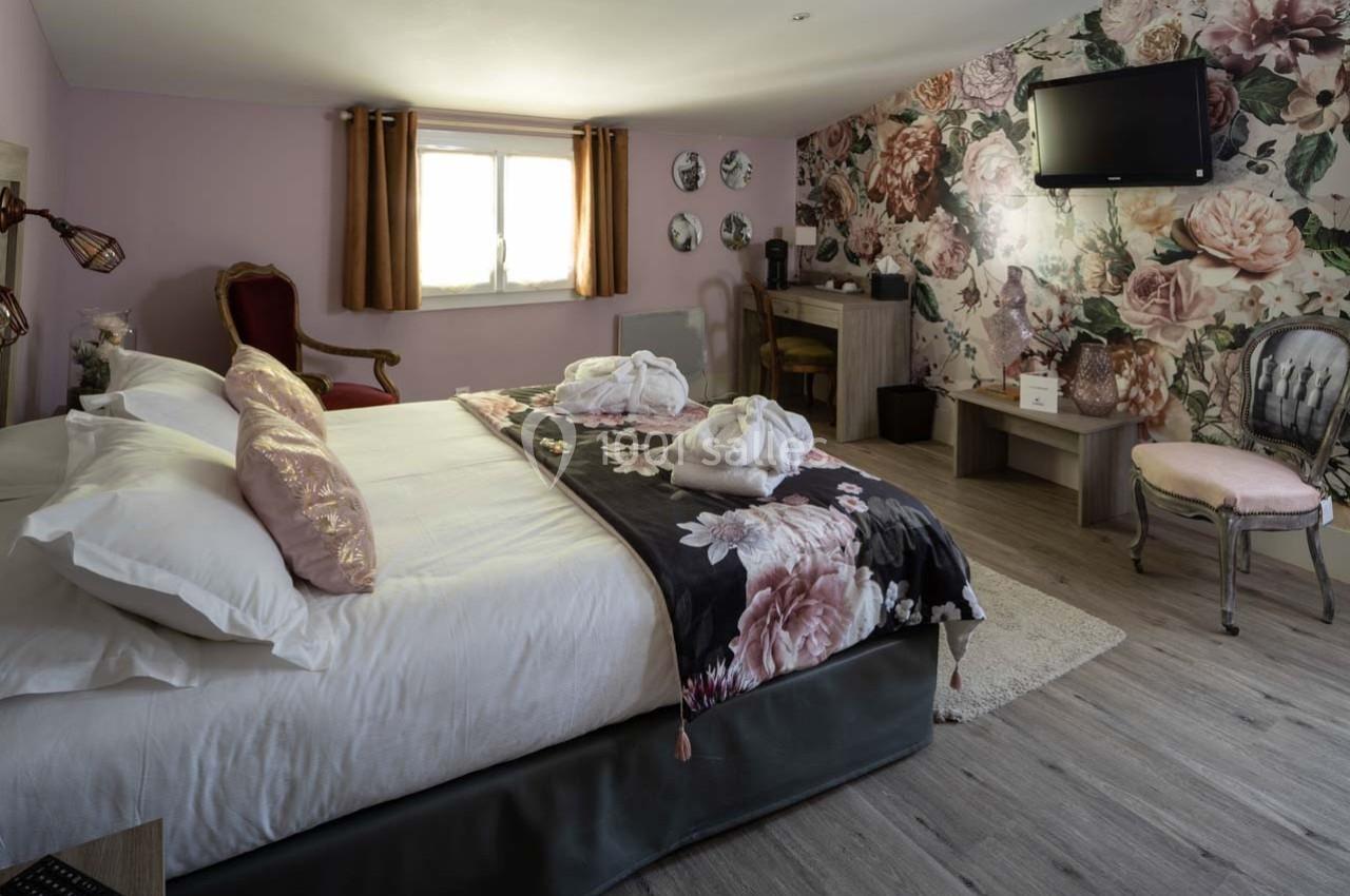 Chambre décorée avec un lit double, fauteuil, bureau, télévision murale et mur fleuri, éclairée par une fenêtre.