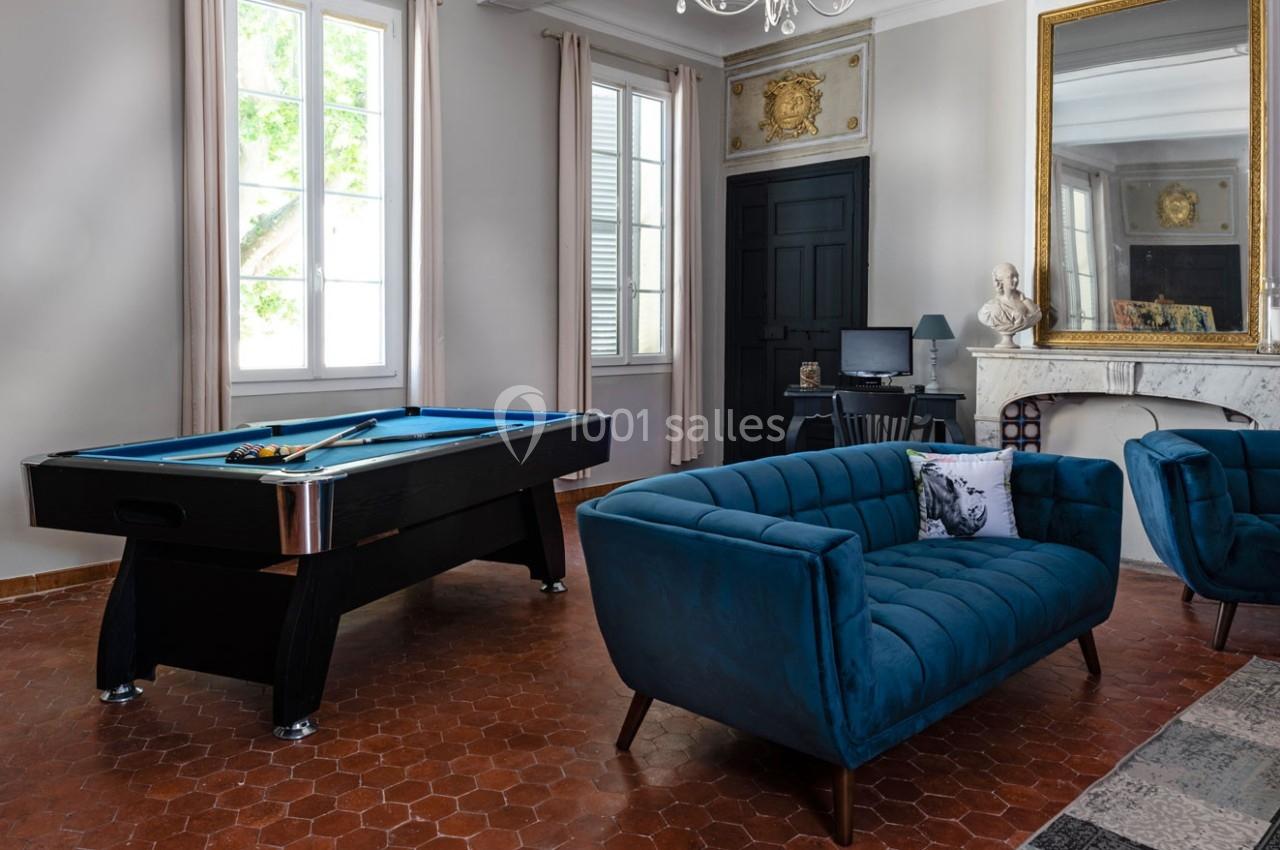 Salle de séjour lumineuse avec un billard, des fauteuils bleus, un bureau, une cheminée et un grand miroir doré.