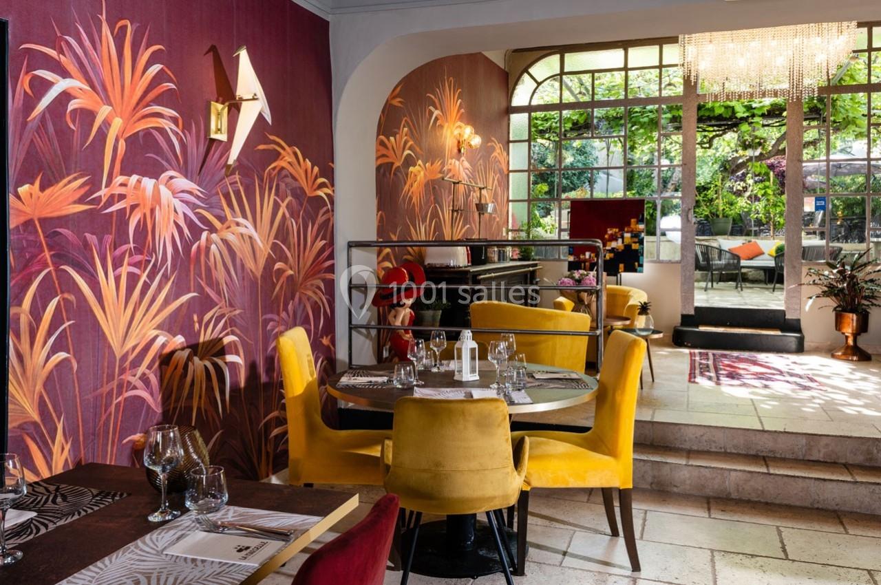 Salle de restaurant élégante avec murs ornés de motifs végétaux colorés, mobilier jaune et vue sur une terrasse verdoyante.