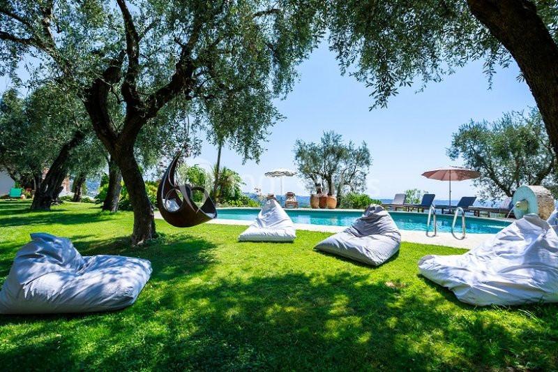 Espace extérieur avec piscine, transats, poufs blancs sur pelouse et arbres offrant de l'ombre.