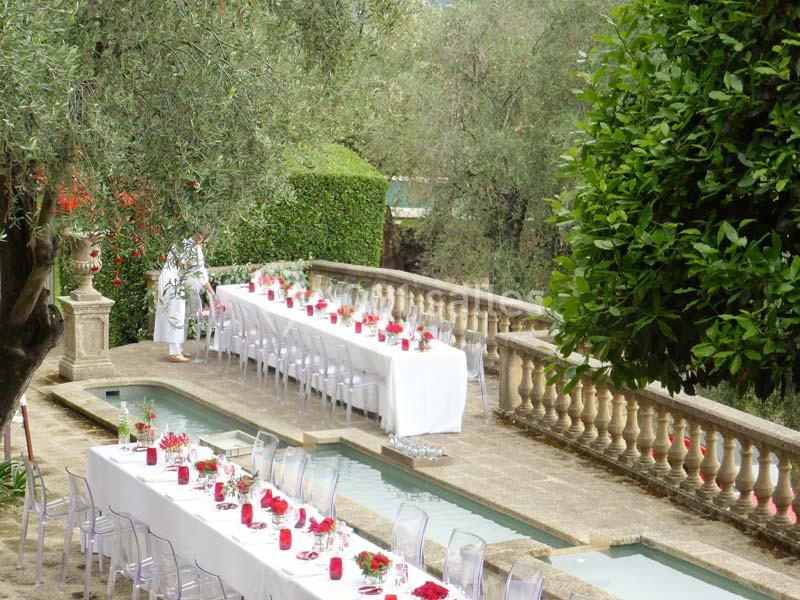 Tables dressées avec nappes blanches et décorations rouges dans un jardin méditerranéen avec fontaine et oliviers.