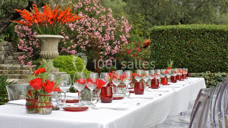 Table dressée en extérieur avec nappes blanches, verres et fleurs rouges, entourée de végétation et de fleurs colorées.