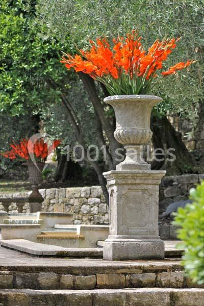 Vase en pierre contenant des fleurs orange vives, placé dans un jardin avec des arbres et des murs en pierre en arrière-plan.