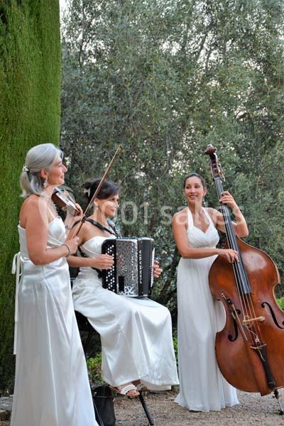 Trois musiciennes en robes blanches jouent du violon, de l'accordéon et de la contrebasse en extérieur près d'arbres.