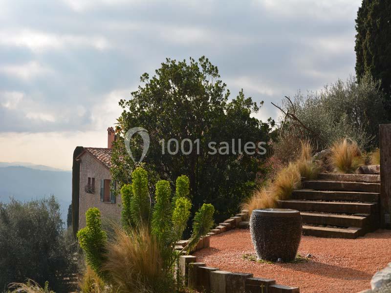 Jardin méditerranéen avec poterie, escaliers en pierre, végétation variée et maison en arrière-plan sous un ciel nuageux.