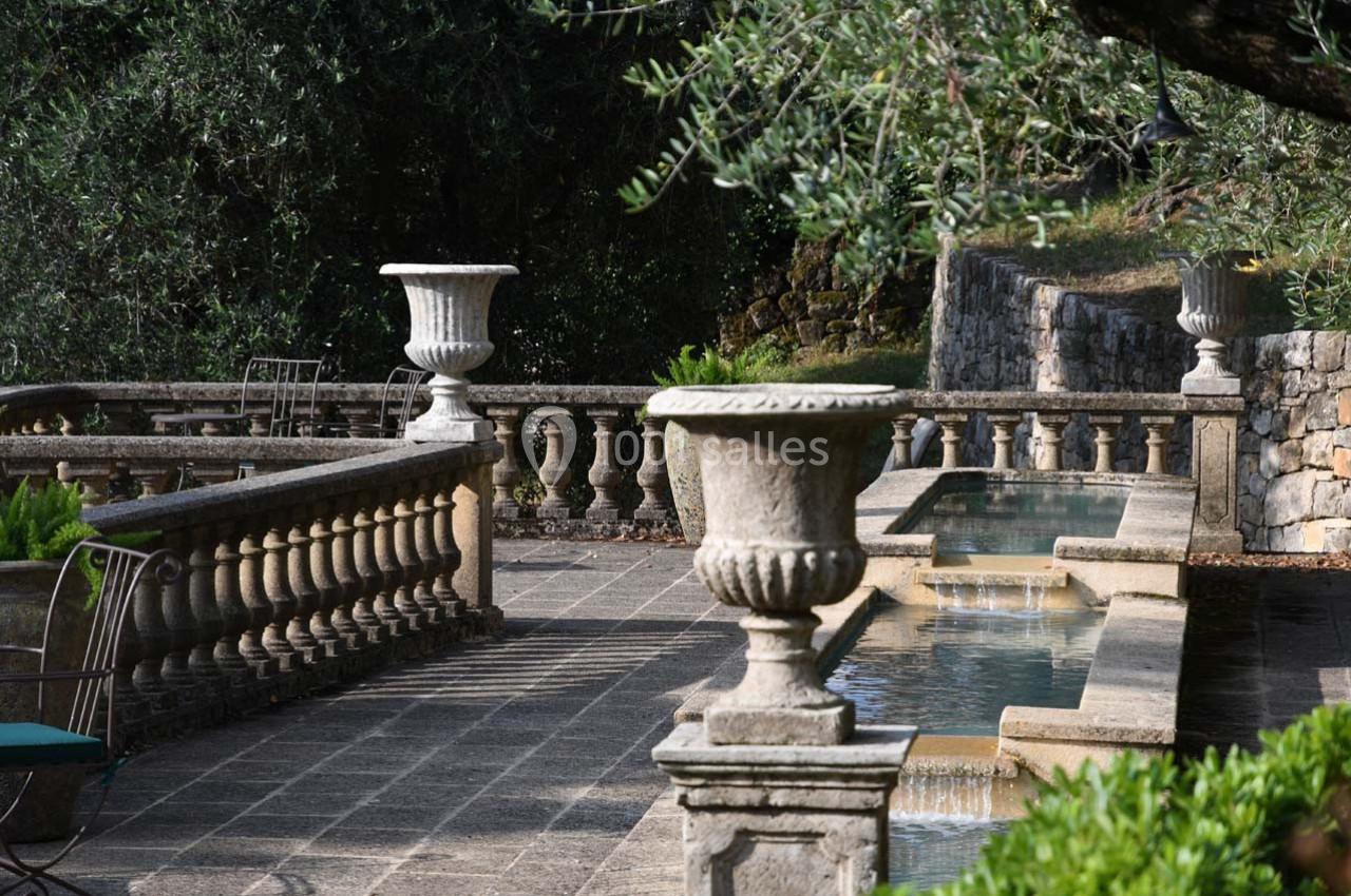 Terrasse en pierre avec balustrades, fontaines en cascade et urnes décoratives entourées de végétation.