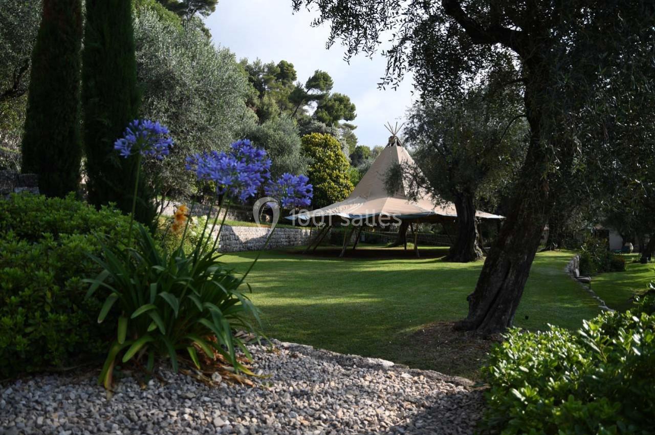 Tente beige installée dans un jardin verdoyant avec des oliviers et des fleurs violettes au premier plan.