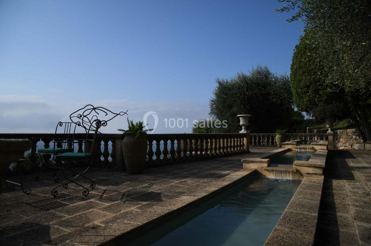 Terrasse en pierre avec bassins d'eau, balustrade surplombant un paysage, chaises en fer forgé et végétation méditerranéenne.
