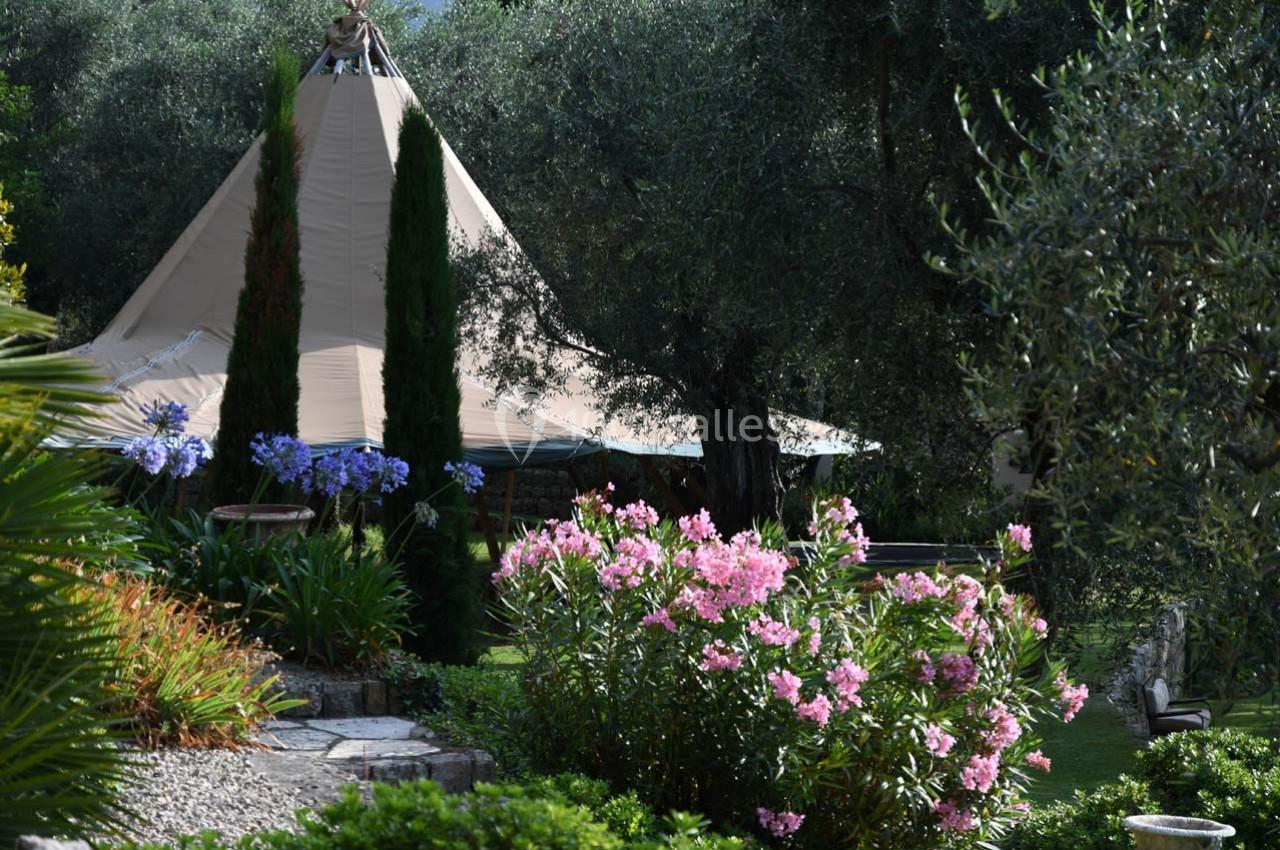 Jardin verdoyant avec fleurs colorées, arbres et une grande tente beige en arrière-plan.