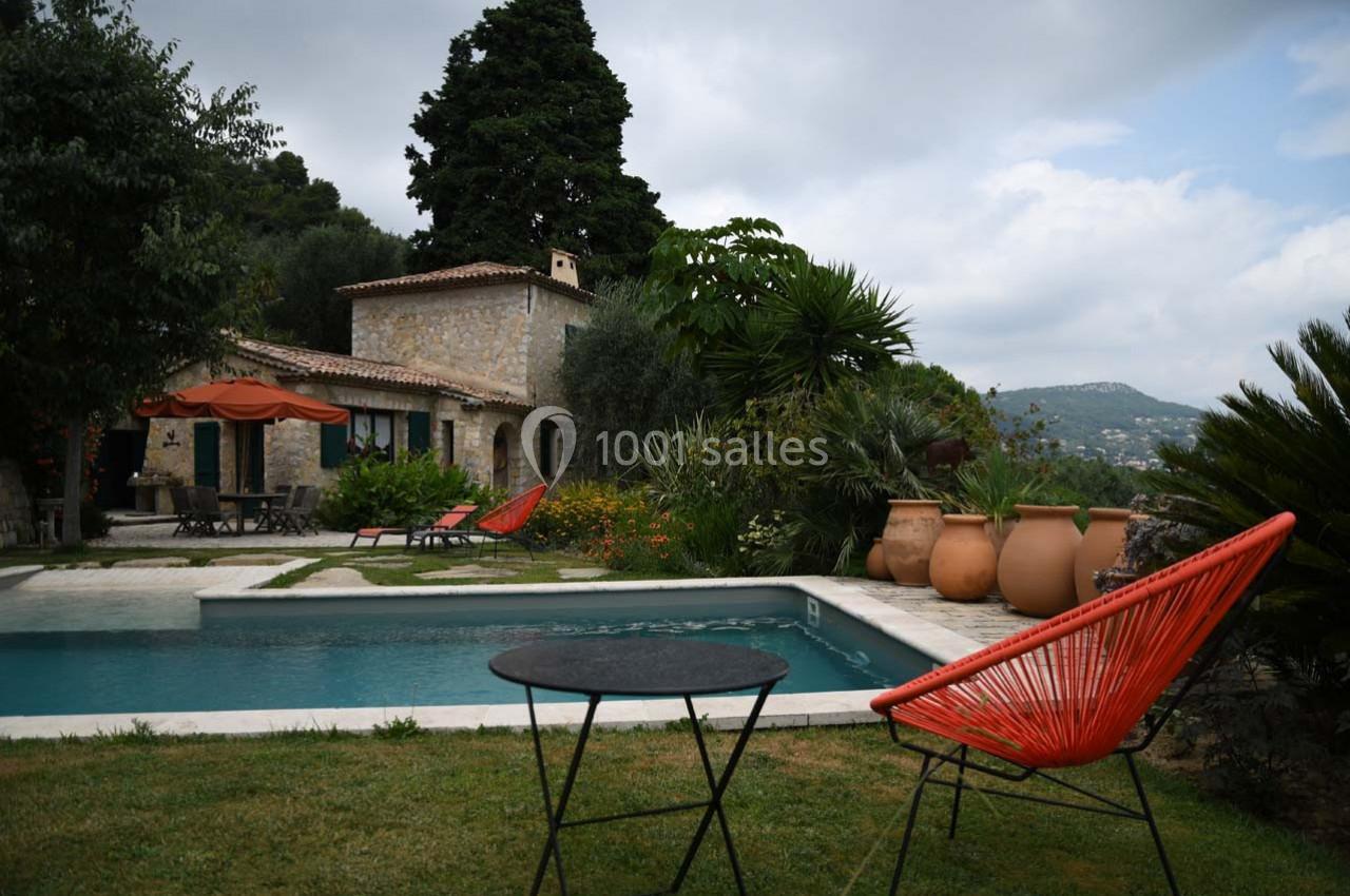 Jardin avec piscine, maison en pierre, chaises colorées et végétation méditerranéenne sous un ciel nuageux.