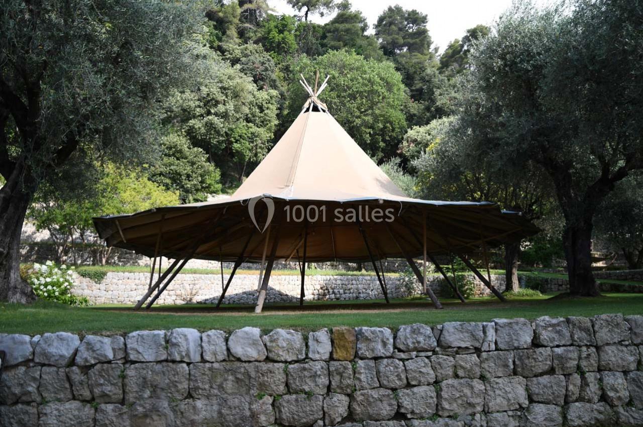 Tente tipi beige installée sur une pelouse, entourée d'arbres et de murets en pierre.