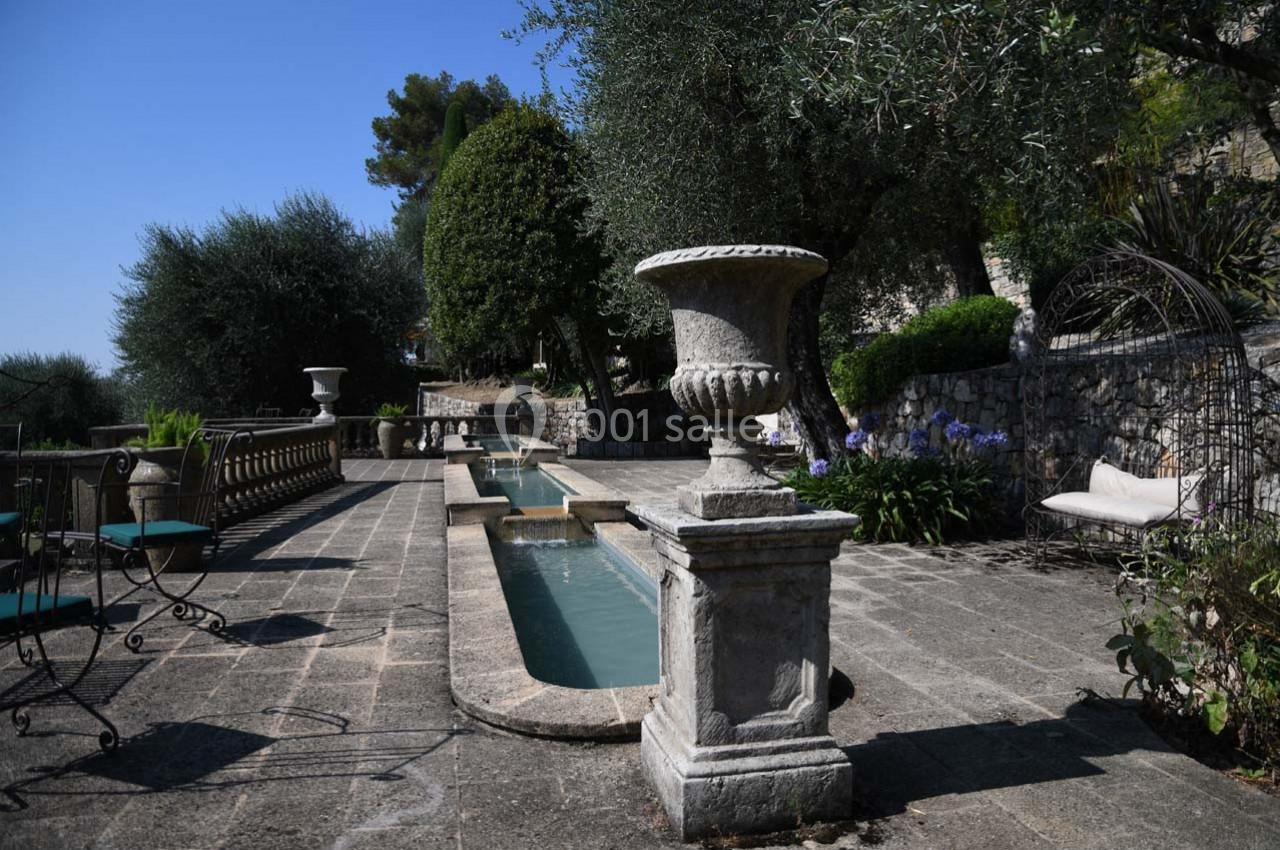 Terrasse en pierre avec bassin central, urne décorative, chaises en fer forgé et végétation méditerranéenne.
