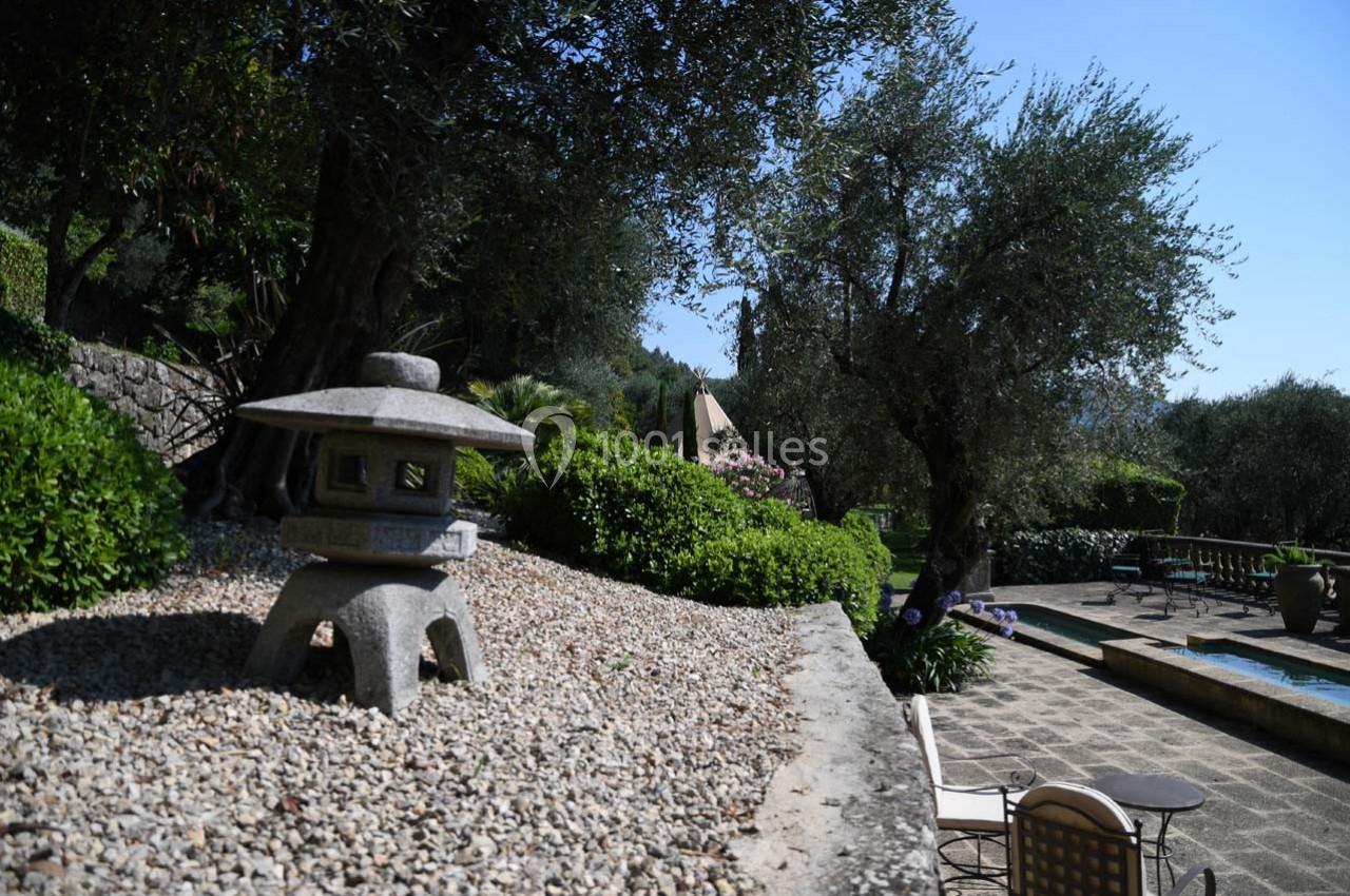 Jardin avec lanterne en pierre, terrasse pavée, piscine et oliviers sous un ciel dégagé.