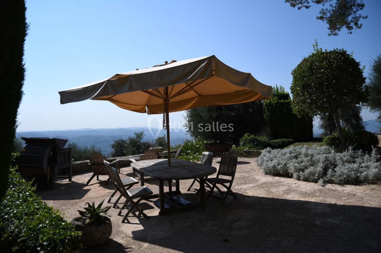 Table en bois entourée de chaises sous un grand parasol beige, située dans un jardin avec vue dégagée sur la campagne.