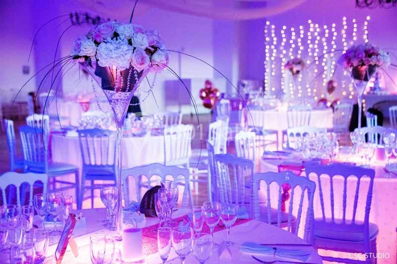 Salle de réception décorée avec des tables dressées, des fleurs en centre de table et un éclairage violet.